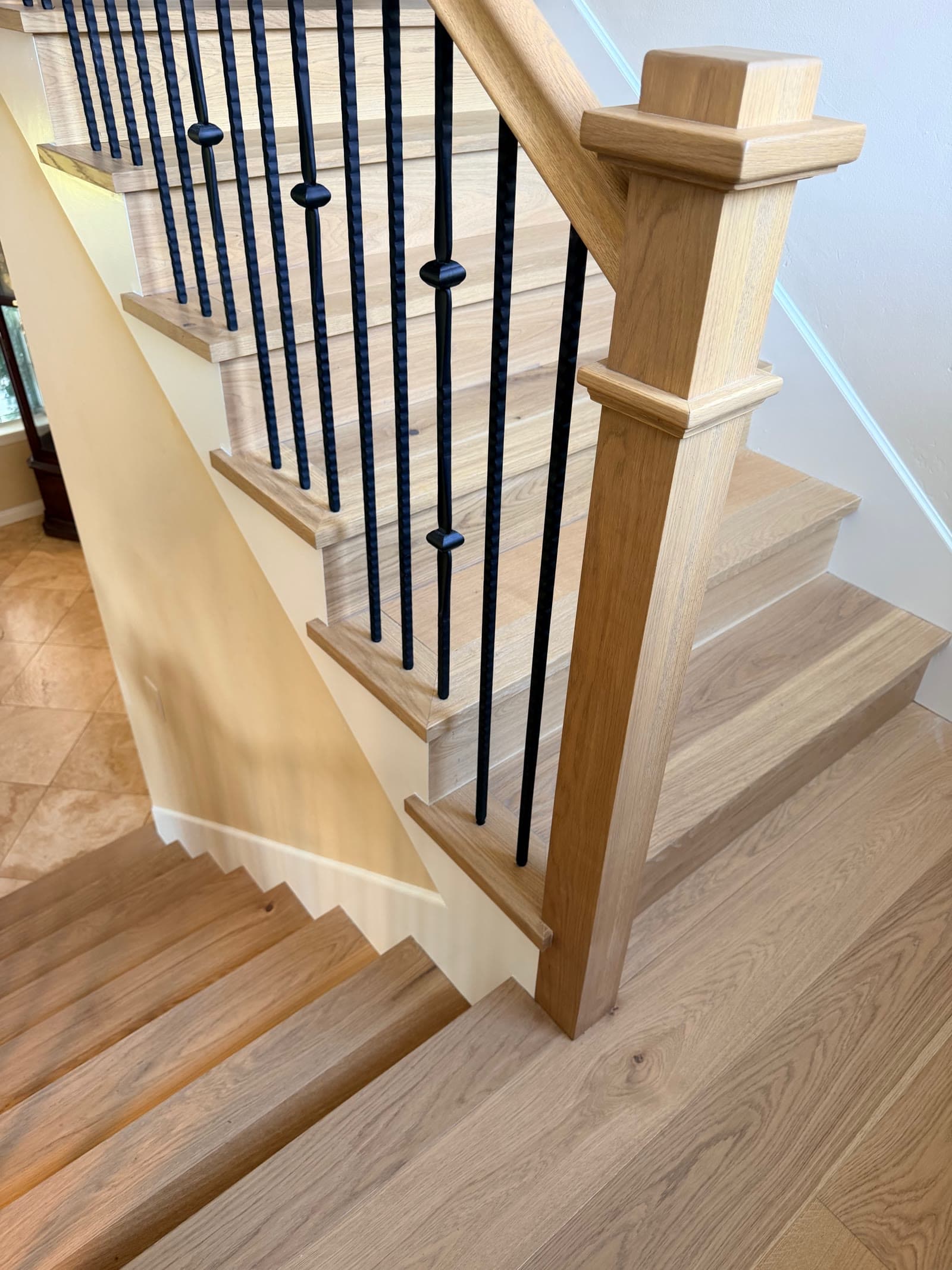 Bottom of a white oak staircase with a square newel post meeting a travertine entry floor